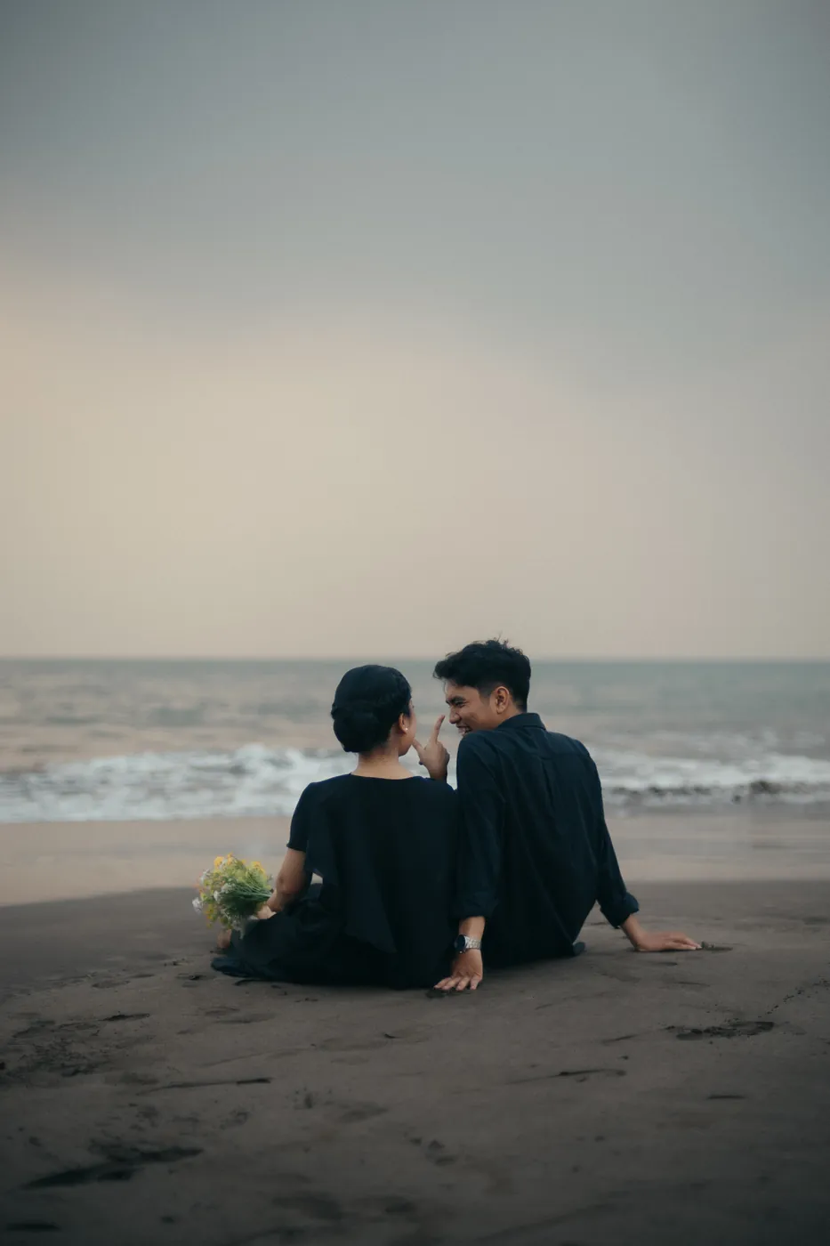 Bastian and Vero sitting together on the sand by the sea, sharing a laugh